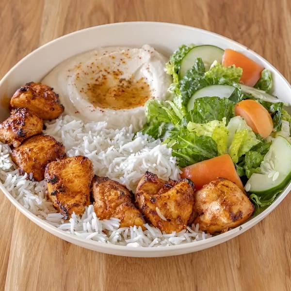 A Mediterranean chicken and rice bowl with hummus and a side salad on a wooden table.
