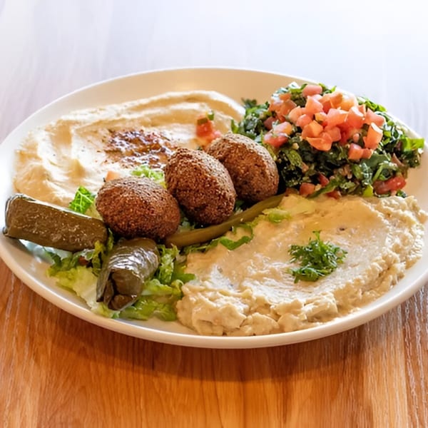 A Mediterranean platter with hummus, falafel, tabbouleh, grape leaves, and pickles on a white plate.